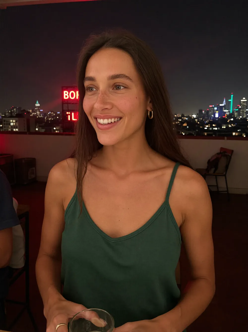 Lina rit sur un toit-terrasse la nuit, haut satiné vert, enseignes au néon et ville en arrière-plan