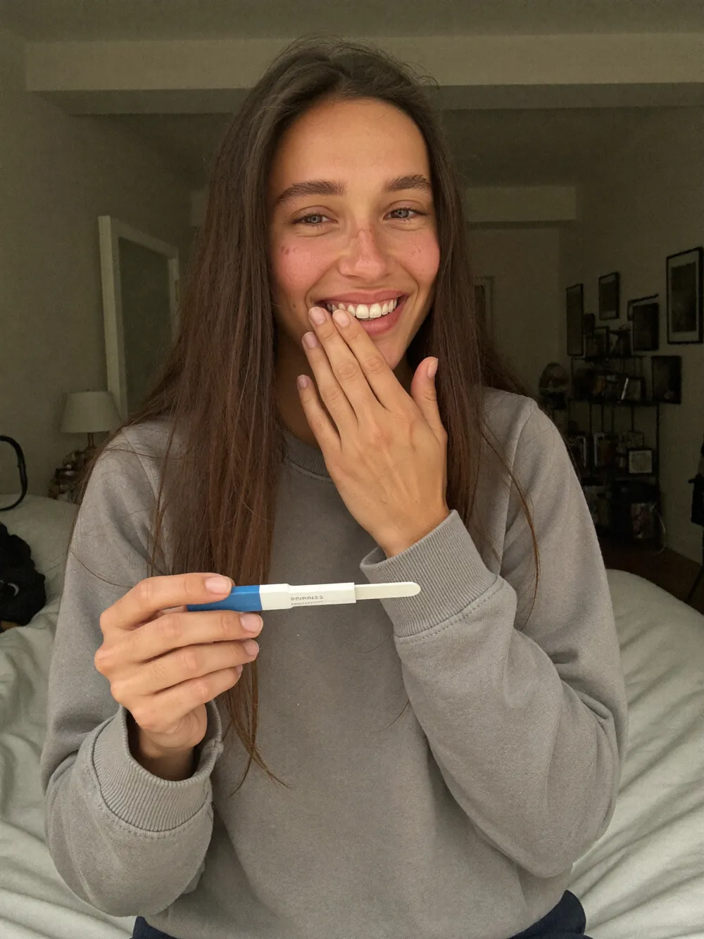 Lina assise sur son lit, un test de grossesse à la main, l'autre main devant la bouche, riant et pleurant à la fois